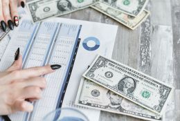 Obrazek dekoracyjny przedstawiający kobiecą dłoń wskazującą na tabelę oraz banknoty dolarów na stole