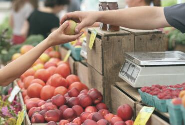Zdjęcie przedstawia stoisko owocowe. Widoczne sa jedynie ręce klienta i sprzedawcy, które przekazują sobie jabłko.