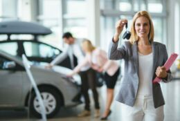Uśmiechnięta kobieta trzymająca klucze do samochodu, w tle samochód i pochyleni kobieta oraz mężczyzna