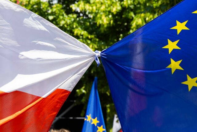 Flaga Polski i Unii Europejskiej związane razem, w tle liście drzewa, słoneczna pogoda.