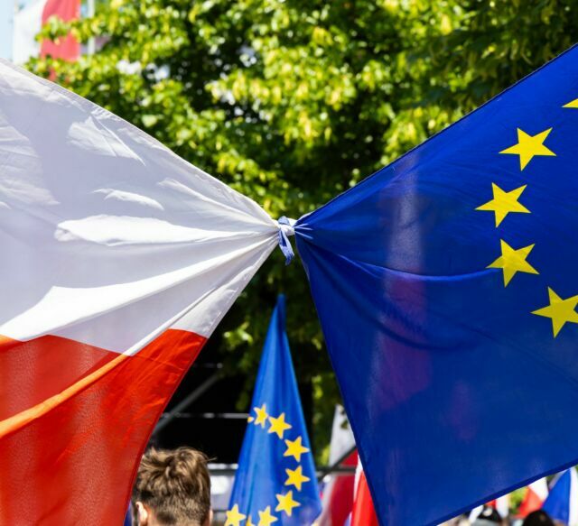 Flags of Poland and the European Union tied together, tree leaves in the background, sunny weather.
