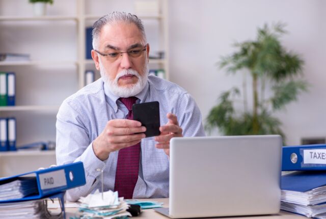 Starszy mężczyzna przed laptopem. W ręku trzyma telefon, patrzy na wyświetlacz