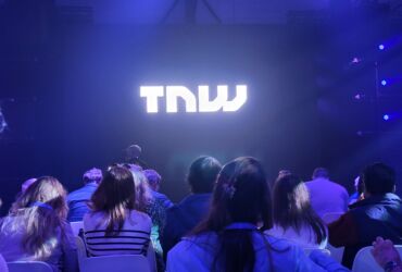 Ludzie siedzący na konferencji. Na ekranie wyświetla się TNW. Przyciemnione światło.