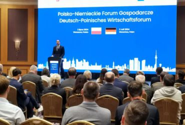 On stage, a man giving a speech. A large screen in the background. In the foreground, conference participants