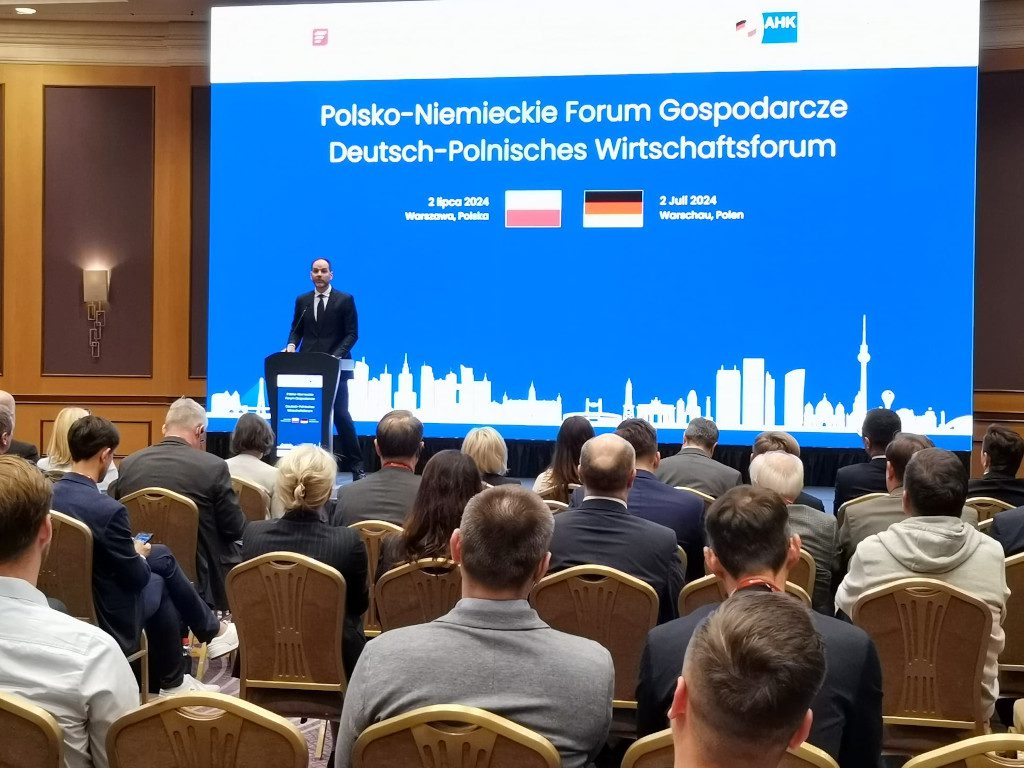 On stage, a man giving a speech. A large screen in the background. In the foreground, conference participants