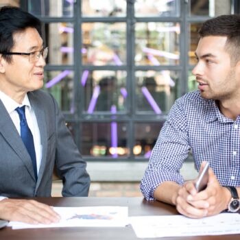 Azjata i Europejczyk prowadzą rozmowę przy stoliku, na który leżą wykresy