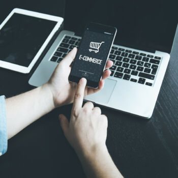 Mężczyzna trzyma w ręku smartfona, na którym widać napis e-commerce. Na biurku także laptop i tablet