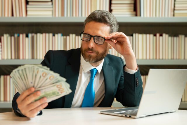Biznesman w okularach siedzi przed laptopem i spogląda na trzymany w ręku wachlarz pieniędzy
