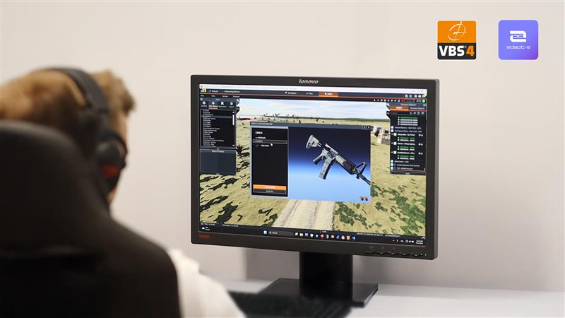 The photo shows a person in front of a monitor displaying a VBS simulation – an image of a weapon and a battlefield